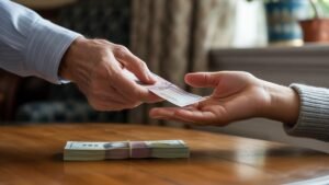 Realus vyresniojo rankos vaizdas, padovanojantis dovaną jaunesnei rankai, subtilią krūvą banknotų, matomų ant šilto medinio stalo. Sutelkite dėmesį į finansinį dosnumą ir šeimos ryšius.