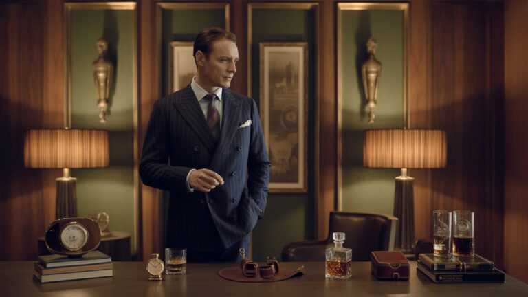 Išmanantis džentelmenas stovi prabangiame, „Art Deco“ įkvėptoje studijoje, žavėdamasi aukščiausios kokybės daiktų kolekcija, pavyzdžiui, senoviniu laikrodžiu, puikiu viskio dekaneriu ir individualiomis odos gaminiais. Fotorealistinis, dramatiškas aplinkos apšvietimas, platus kadras.